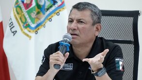 Hombre serio hablando ante un micrófono con la mano estirada.