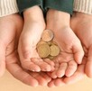 Manos de un adulto sosteniendo las manos de un niño que sostiene un puño de euros con una mesa de madera de fondo
