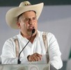 Carlos Alberto Manzo Rodríguez hablando por un micrófono ante un podio con una bandera de México difuminada en el fondo durante presentación pública