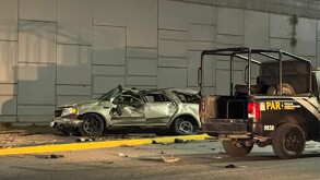 Se observa un carro color gris destruido  y una patrulla a un lado; es de noche.