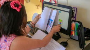 Niña vista de espaldas sosteniendo un libro delante de la pantalla de una computadora con cuadernos y plumones de colores alrededor