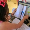 Niña vista de espaldas sosteniendo un libro delante de la pantalla de una computadora con cuadernos y plumones de colores alrededor