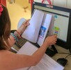 Niña vista de espaldas sosteniendo un libro delante de la pantalla de una computadora con cuadernos y plumones de colores alrededor