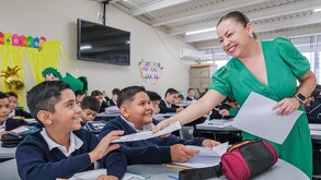Maestra de Jalisco sonriendo mientras entrega un examen a dos estudiantes con otros varios alumnos desenfocados en el fondo dentro de un salón de clases