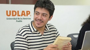 Alumno con un libro en las manos frente a una laptop con logo de la UDLAP atrás
