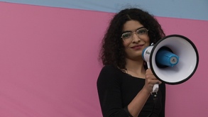 Mujer trans con un megáfono en la mano. Al fondo, una bandera rosa.