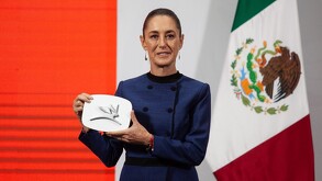 Mujer sosteniendo un objeto rectangular junto a una bandera de México.
