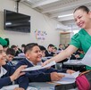 Maestra de Jalisco sonriendo mientras entrega un examen a dos estudiantes con otros varios alumnos desenfocados en el fondo dentro de un salón de clases