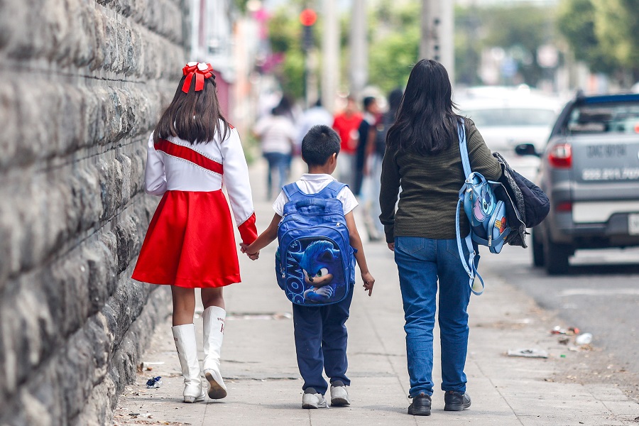 Dos niños con uniforme escolar caminan junto a su mamá.