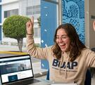 Estudiante festeja con material de estudio en su mesa y detrás las instalaciones de la BUAP