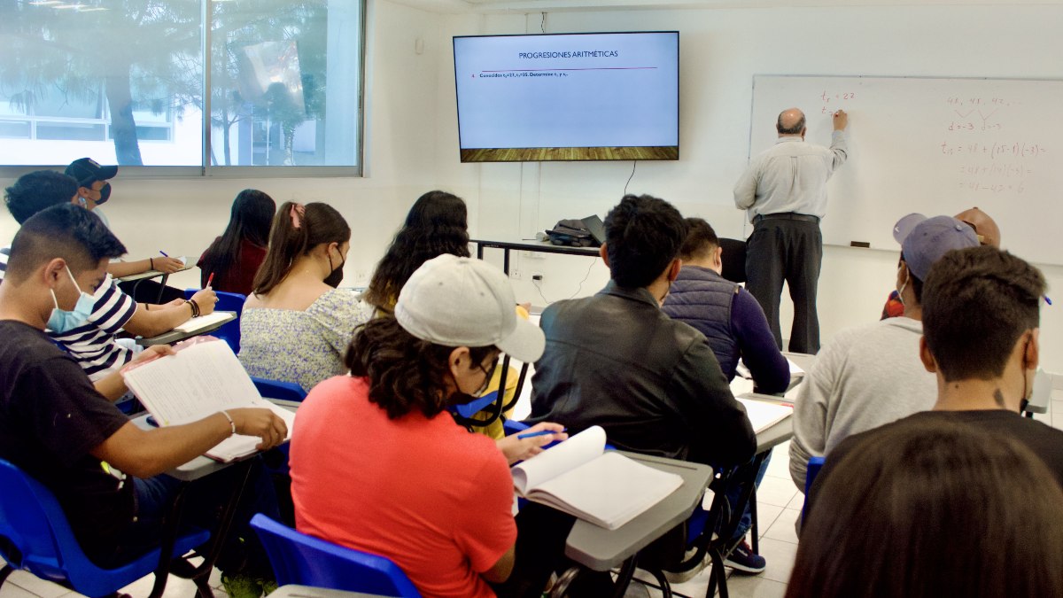 Estudiantes realizan apuntes durante una clase y el profesor anota en el pizarrón dentro de un aula de la BUAP
