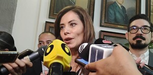 Verónica Delgadillo hablando con múltiples micrófonos de frente con personas detrás de ella y diversos cuadros en el fondo
