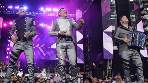 Se observa a tres hombres en un escenario y uno de ellos cantando.
