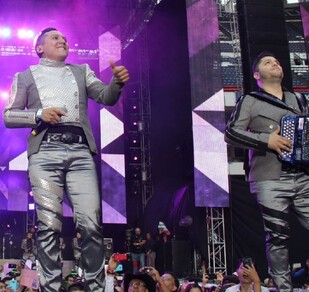 Se observa a tres hombres en un escenario y uno de ellos cantando.