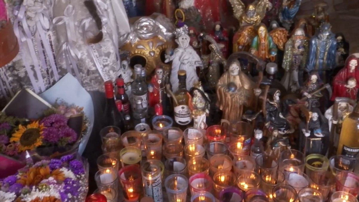 Figuras pequeñas de la Santa Muerte apoyadas en el suelo rodeadas de veladoras, flores, botellas de tequila y demás ofrendas