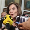 Verónica Delgadillo hablando con múltiples micrófonos de frente con personas detrás de ella y diversos cuadros en el fondo