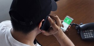 Hombre visto desde arriba vistiendo una gorra negra y sosteniendo una llamada telefónica con un celular en cada mano y un teléfono fijo al frente en una mesa