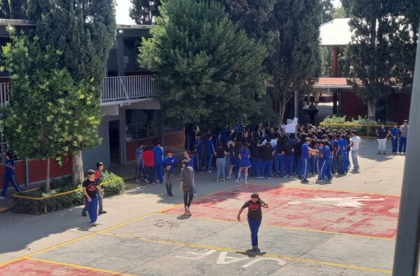Se observa a varios jóvenes que están parados en un patio de una escuela; traen uniforme.