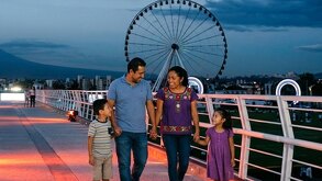 Familia camina por un puente y de fondo la emblemática rueda de Puebla