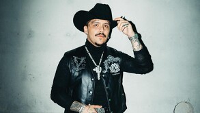 Cantante Christian Nodal parado delante de una pared blanca viendo a la cámara mientras sostiene su sombrero con la mano izquierda