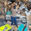 Hombre visto de espaldas parado delante de la cochera de una casa llena de basura, cajas de cartón y un auto de modelo antiguo