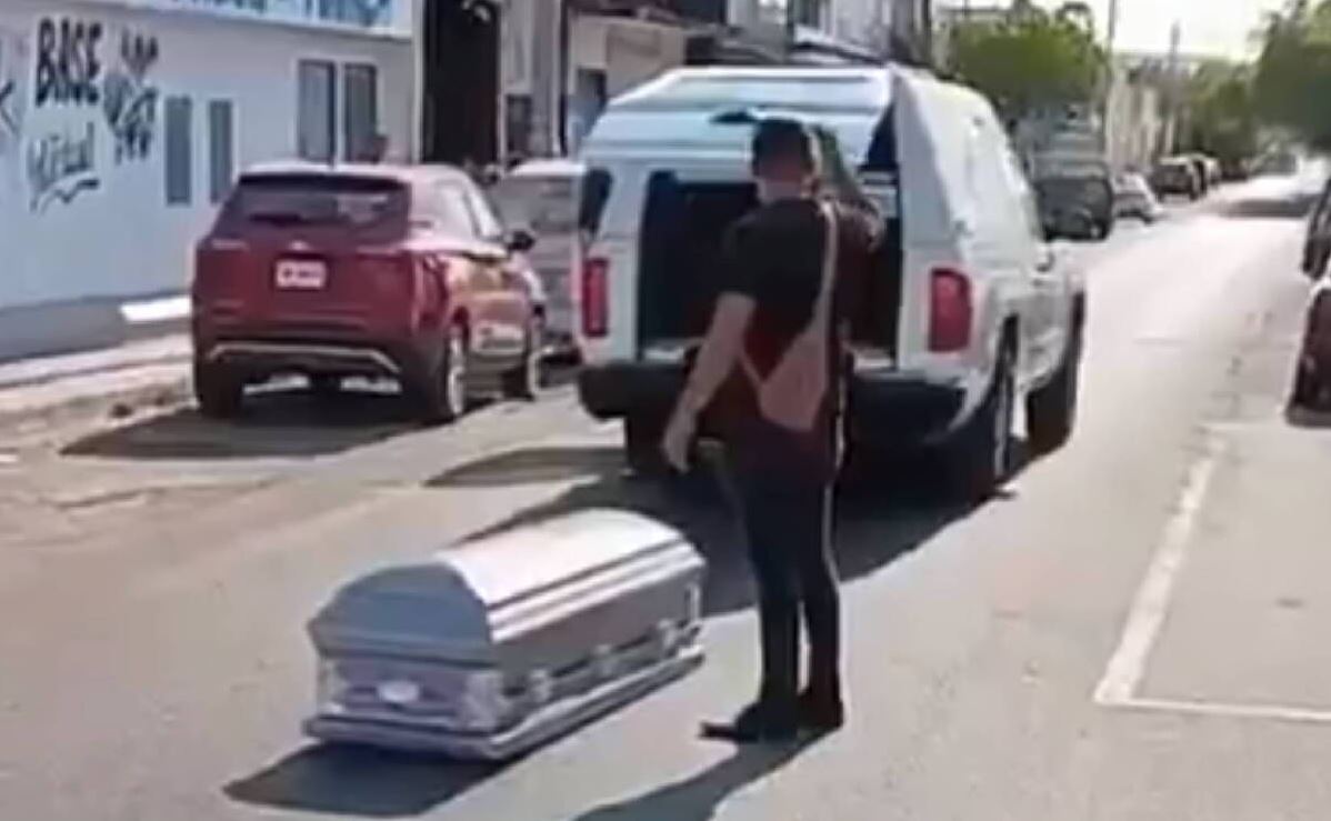Ataúd en medio de la calle con un hombre visto de espaldas a un lado y una carroza funeraria con las puertas abiertas más adelante