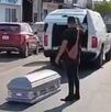 Ataúd en medio de la calle con un hombre visto de espaldas a un lado y una carroza funeraria con las puertas abiertas más adelante