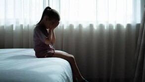 Niña sentada en el borde de una cama tapándose la cara con las manos delante de una ventana