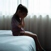 Niña sentada en el borde de una cama tapándose la cara con las manos delante de una ventana