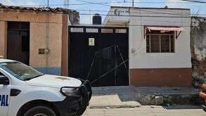 Exterior de una casa con portón negro acordonada por una cinta policiaca amarilla y una patrilla de la Policía de Zapotiltic estacionada enfrente