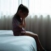 Niña sentada en el borde de una cama tapándose la cara con las manos delante de una ventana