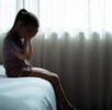 Niña sentada en el borde de una cama tapándose la cara con las manos delante de una ventana