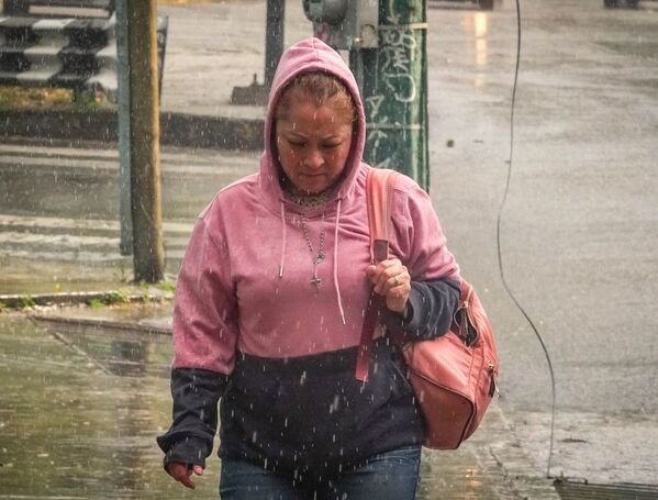 Una mujer camina bajo la lluvia en una zona urbana, vistiendo sudadera con capucha y cargando un bolso, mientras el pavimento mojado refleja las condiciones cli