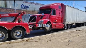 Tráiler blanco con cabina roja siendo remolcado por una grúa al lado de un camino de tierra y un muro de contención en Tlaquepaque
