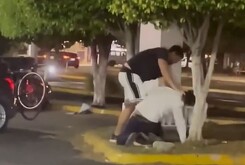 Un hombre agarrando por el cuello de la camisa a otro que se encuentra en el suelo de la jardinera de un estacionamiento