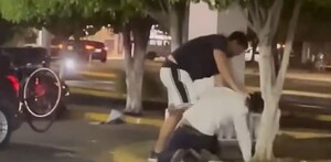 Un hombre agarrando por el cuello de la camisa a otro que se encuentra en el suelo de la jardinera de un estacionamiento