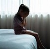 Niña sentada en el borde de una cama tapándose la cara con las manos delante de una ventana