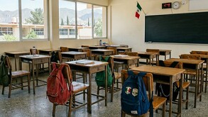 Se observa un salón de clases, hay pupitres solos, mochilas, un pizarrón y un reloj.