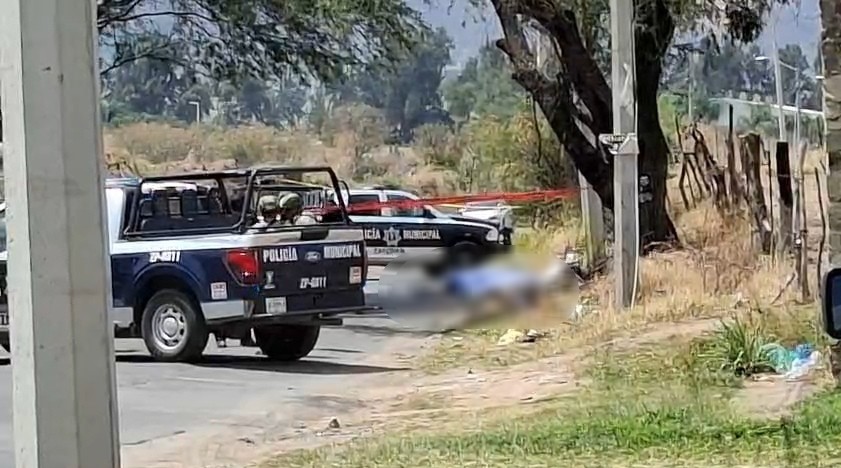 Patrullas de la Policía de Zapopan en un área acordonada con una cinta policiaca roja donde hay un cuerpo cubierto por una manta azul blureado