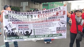 Se observa a personas de pie con una pancarta en las manos, visten playera roja.