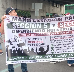 Se observa a personas de pie con una pancarta en las manos, visten playera roja.