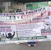 Se observa a personas de pie con una pancarta en las manos, visten playera roja.
