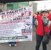 Se observa a personas de pie con una pancarta en las manos, visten playera roja.