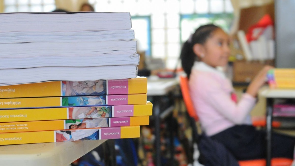 Se observa a una niña sentada en un pupitre, y hay unos libros sobre una mesa.
