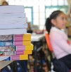 Se observa a una niña sentada en un pupitre, y hay unos libros sobre una mesa.