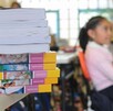 Se observa a una niña sentada en un pupitre, y hay unos libros sobre una mesa.