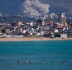 Unas personas nadan en una playa pública mientras al fondo se ve humo tras un ataque aéreo israelí que impactó la aldea de Qlaileh, vista desde Tiro, Libano
