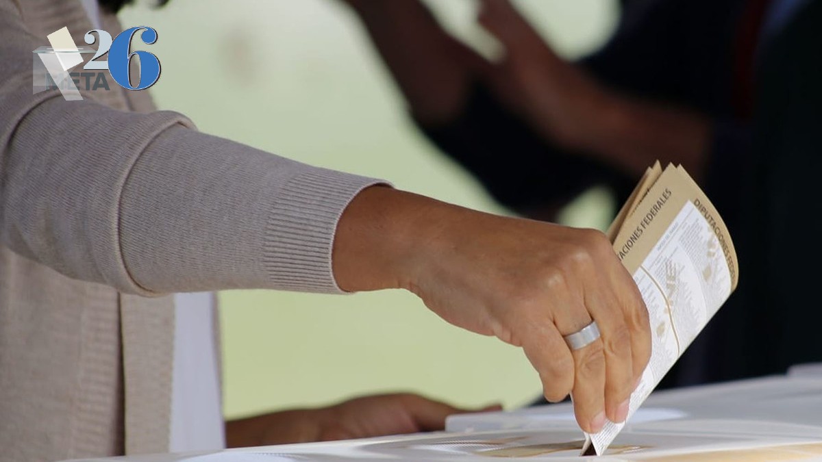 Imagen en primer plano de una mano colocando una boleta electoral dentro de una urna. El fondo es desenfocado.