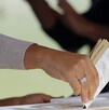 Imagen en primer plano de una mano colocando una boleta electoral dentro de una urna. El fondo es desenfocado.