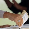 Imagen en primer plano de una mano colocando una boleta electoral dentro de una urna. El fondo es desenfocado.